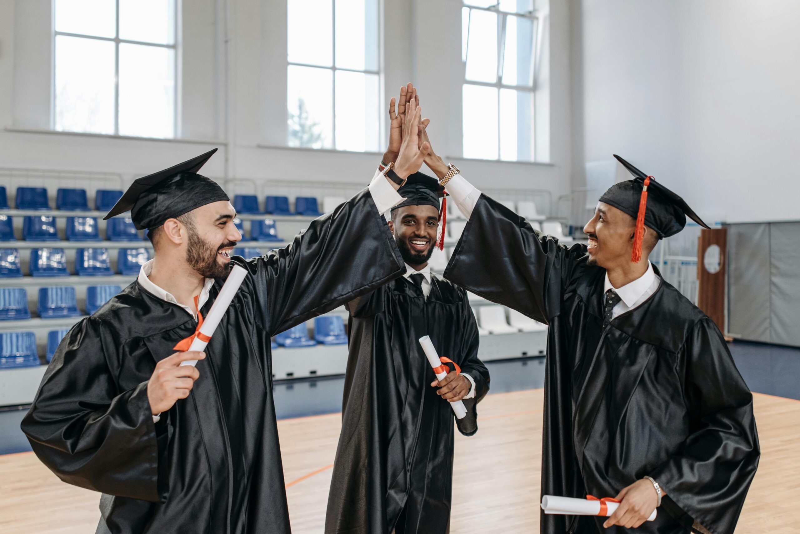 Three graduates in gowns celebrating their success with high fives indoors.